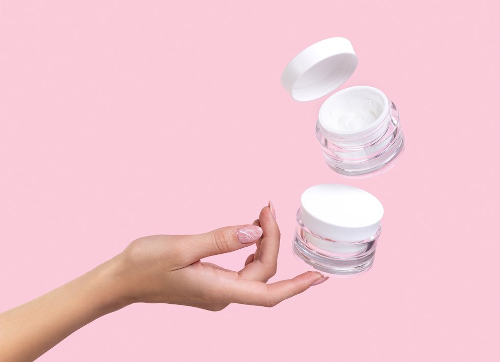 Open jar with cosmetic cream with womans hand on pink background