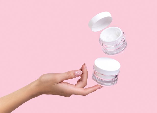 Open jar with cosmetic cream with womans hand on pink background